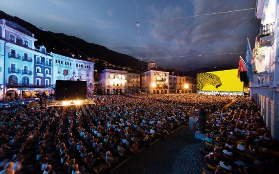 Auf der großen Piazza in Locarno stehen zwischen den historischen Gebäuden unzählige Stühle mit Personen und eine große Leinwand ist aufgebaut. 