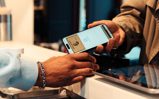 Eine Person bezahlt kontaktlos per Smartphone mit goldener Kreditkarte auf dem Display an einem Kartenlesegerät auf einem Tresen.