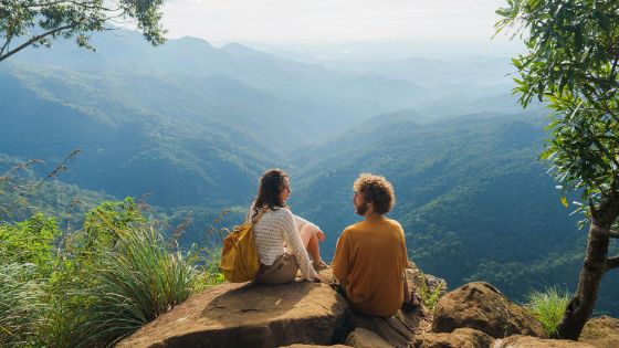 Eine Frau und ein Mann sitzen auf einem kleinen Hügel und schauen auf eine Berglandschaft. 