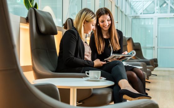 Zwei stilvoll gekleidete Frauen sitzen in grossen Ledersesseln und blicken gemeinsam in eine Zeitschrift, auf einem Tisch daneben steht eine weisse Tasse.