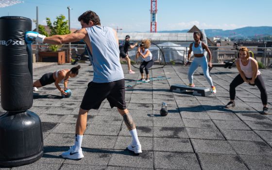 Mehrere Personen trainieren mit unterschiedlichen Fitnessgeräten auf einer Dachterrasse bei Sonnenschein.