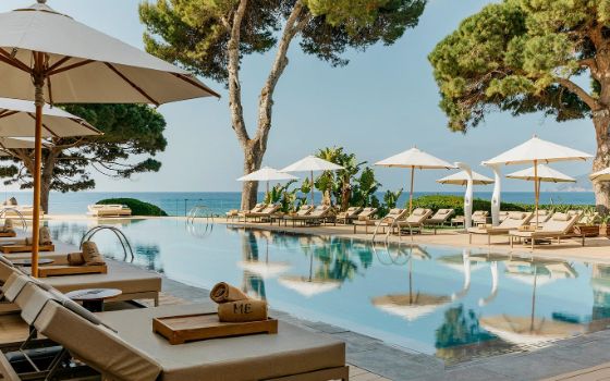 An einem Hotelpool stehen Liegen und Sonnenschirme, im Hintergrund liegt das Meer. 