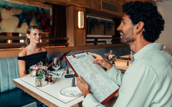 Eine Frau in schwarzem Kleid und ein Mann im Hemd sitzen sich gegenüber mit Speisekarten in den Händen an einem Tisch in einem stilvoll eingerichteten Restaurant. 