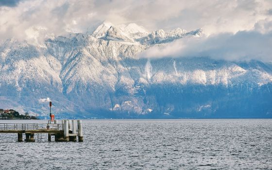 Schneebedeckte Alpen hinter dem Genfersee mit Steg im Vordergrund und dramatischer Wolkenstimmung im Winter.