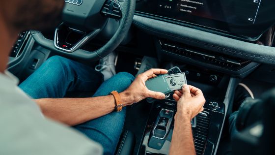 Ein Mann am Steuer eines modernen Autos der Marke Audi zieht eine silberne Kreditkarte von American Express aus einem Lederetui an einem Smartphone über der Mittelkonsole.