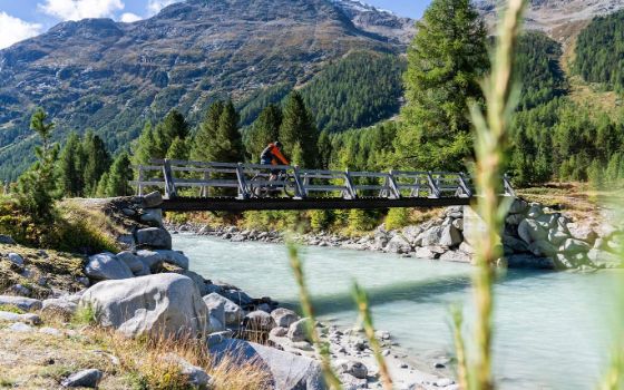 Ein Radfahrer fährt über eine Brücke, die über einen Bach führt, und umgeben ist von Bergen und Natur. 