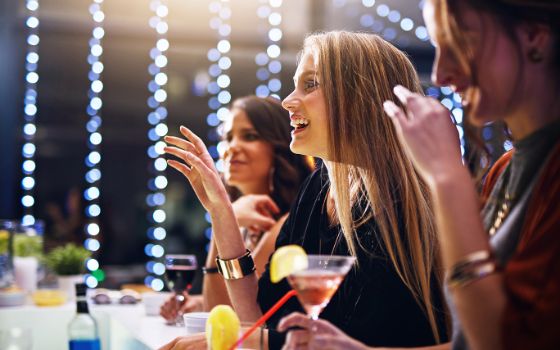 Eine junge Frau steht an einer Bar und hebt eine Hand, um ein Getränk zu bestellen.