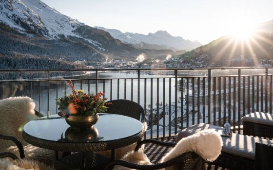 Terrasse mit Tisch und Sitzgelegenheiten an einem See vor schneebedecktem Bergpanorama im tiefstehenden Sonnenlicht.