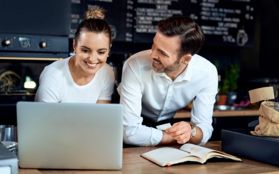 Junge Frau in weißem Oberteil blickt lächelnd auf einen Laptop, neben ihr ein Mann im weißen Hemd, er schaut sie lächelnd an, hat vor sich ein Buch liegen. Im Hintergrund eine schwarze Tafel mit weißer Schrift.