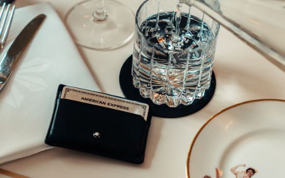 Eine silberne Kreditkarte von American Express in einem schwarzen Lederetui liegt neben einem Wasserglas auf einem eingedeckten Tisch. 