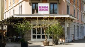 Aussenaufnahme eines Restaurants mit der Aufschrift BUNZAI in violetten Lettern über dem Eingang an einer Strassenecke mit Bäumen in Kübeln.