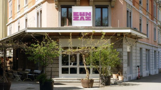 Aussenaufnahme eines Restaurants mit der Aufschrift BUNZAI in violetten Lettern über dem Eingang an einer Strassenecke mit Bäumen in Kübeln.