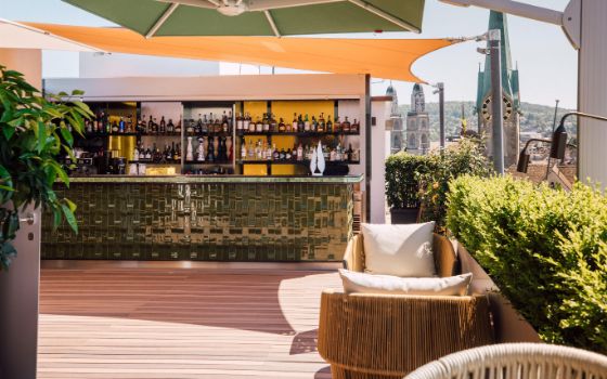 Ein menschenleerer Barbereich auf einer Dachterrasse mit Sitzgelegenheiten vor der Skyline Zürichs bei Sonnenschein.