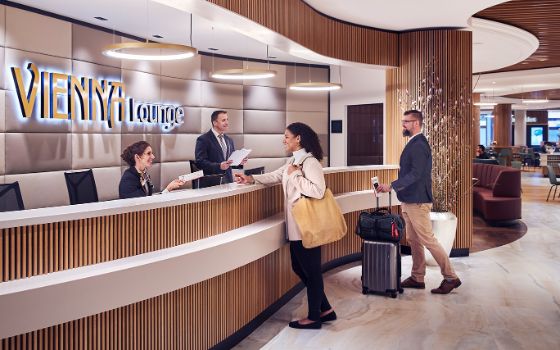 Personen mit Reisegepäck stehen an einer geschwungen Empfangstheke einer modernen Flughafen-Lounge mit Mitarbeitern vor einer Wand mit einem beleuchteten Vienna Lounge Schriftzug.