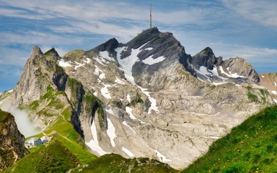 Blick auf die Gipfelstation auf dem Säntis, im Hintergrund ein blauer Himmel. Auf dem Berg liegt Schnee, unten grüne Wiesen. 