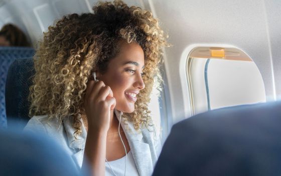 Eine lächelnde Frau mit Kopfhörern sitzt am Fenster in einem Flugzeug.
