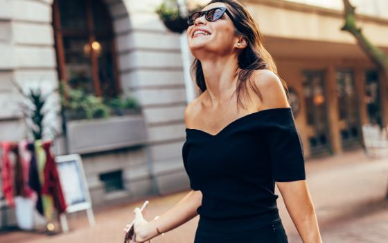 Eine Frau mit schwarzem Kleid und Sonnenbrille spaziert lächelnd in einer Strasse mit Geschäften. 