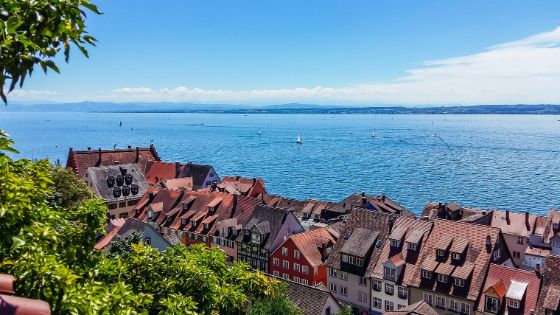 Blick auf historische Giebelhäuser einer Kleinstadt an einem grossen See mit Segelboten von einer bepflanzten Anhöhe aus. 