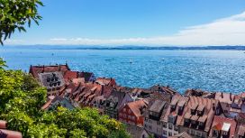 Blick auf historische Giebelhäuser einer Kleinstadt an einem grossen See mit Segelboten von einer bepflanzten Anhöhe aus. 