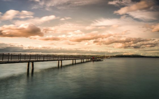 Langer Holzsteg am Bodensee bei Abendstimmung, sanfte Wellen und dramatische Wolkenformationen am Himmel.