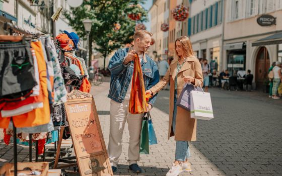 Ein Mann und eine Frau mit Einkaufstüten begutachten ein Kleidungsstück an einem Strassenstand in einer Fussgängerzone bei Sonnenschein. 