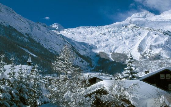 Vor schneebedeckten Bergen stehen Tannen und Häuser, auch sie sind mit viel Schnee bedeckt. 