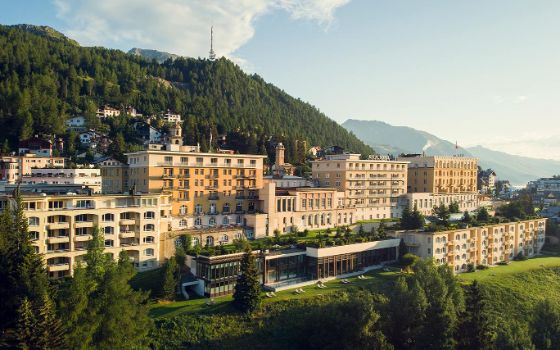 Auf einem Berg steht ein grosses Hotel mit gelber Fassade, umgeben von Bergen und Natur. 