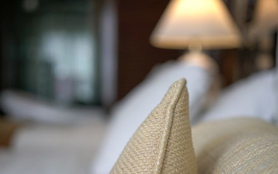 Die Spitze von einem beigen Kissen liegt auf einem Bett, im Hintergrund sind unscharf die Umrisse eines Hotelzimmers wie eine Lampe, Bett und Schrank erkennbar. 