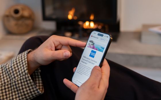 Nahaufnahme von zwei Händen, die ein Smartphone halten, auf dessen Display eine Website mit einem Reisportal mit einem American Express Logo zu sehen ist, vor einem Kamin in einem Wohnzimmer. 