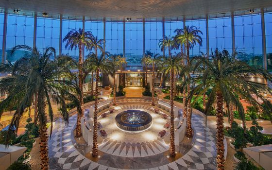 Prunkvolle, beleuchtete Hotellobby im orientalischen Design mit Glasfront bei Dunkelheit. Im Zentrum befindet sich ein kreisrunder Loungebereich aus Marmor, umgeben von zahlreichen Palmen. 