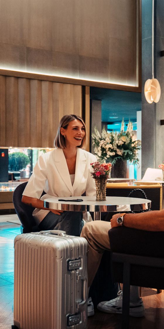 Eine Frau und ein Mann sitzen in einer eleganten Hotel-Lobby an einem Tisch, vor ihnen steht ein schicker Rollkoffer.