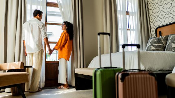 Ein Mann und eine Frau stehen Hand in Hand an einem Fenster in einem modernen, hellen Hotelzimmer, im Vordergrund stehen zwei Hartschalenkoffer. 