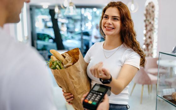 Eine junge Frau steht mit einer Einkaufstüte im Arm in einem Café und hält lächelnd die Smartwatch an ihrem Handgelenk an ein Kartenlesegerät, das eine Person ihr hinhält. 