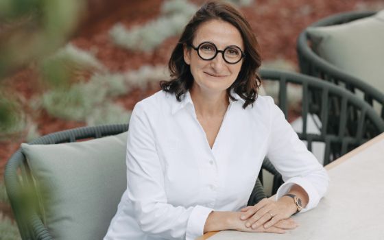 Anne-Sophie Pic trägt eine weisse Bluse und eine Statement-Brille, sie sitzt auf einem Korbstuhl und schaut lächelnd in die Kamera. 