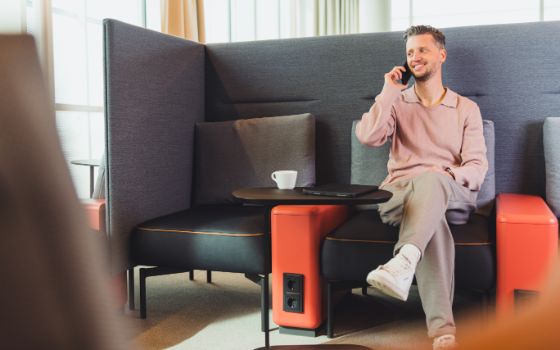 Ein Mann sitzt in einem Sessel mit grosser Rückenlehne und Steckdosen an einem Tisch mit einer Tasse und Tablet und telefoniert.