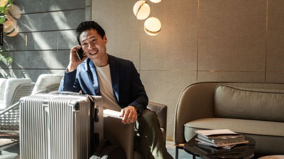 Ein elegant gekleideter Mann sitzt auf einem Sessel in einer Lounge und telefoniert mit seinem Smartphone, vor ihm ein silberfarbener Hartschalenkoffer.