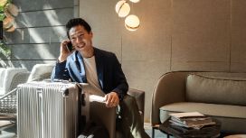 Ein elegant gekleideter Mann sitzt auf einem Sessel in einer Lounge und telefoniert mit seinem Smartphone, vor ihm ein silberfarbener Hartschalenkoffer.
