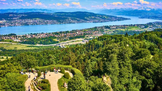 Personen auf einer Aussichtsplattform auf einem bewaldeten Berg mit Blick auf den Zürichsee bei Sonnenschein.