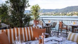 Eine Dachterrasse mit Blick auf den Zürichsee und Berge, die mit Pflanzen dekoriert ist und auf der sich Sofas und Stühle an gedeckten Tischen befinden.