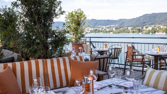Eine Dachterrasse mit Blick auf den Zürichsee und Berge, die mit Pflanzen dekoriert ist und auf der sich Sofas und Stühle an gedeckten Tischen befinden.
