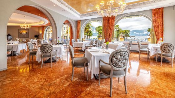 Heller, opulenter Speisesaal mit weiss eingedeckten Tischen auf einem glänzenden, bernsteinfarbenen Boden vor gerundeten Panoramafenstern mit Seeblick. 