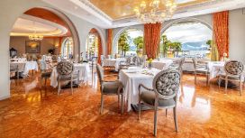 Heller, opulenter Speisesaal mit weiss eingedeckten Tischen auf einem glänzenden, bernsteinfarbenen Boden vor gerundeten Panoramafenstern mit Seeblick. 