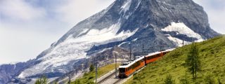 Eine rote Bergbahn fährt auf einer Trasse über einen grasbewachsenen Bergkamm vor der Kulisse eines schneebedeckten Berggipfels.