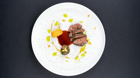 Auf einem weissen Teller liegen vier kleine Stücke von gebratenem Fleisch mit einem Klecks brauner Sosse und einer Krokette.