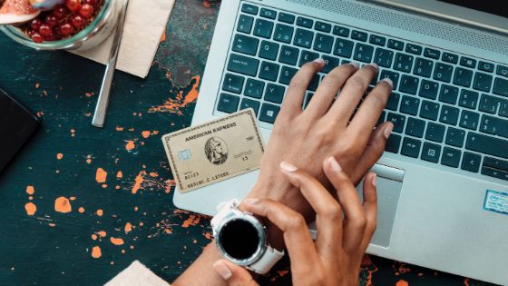 Weibliche Hände über Laptop Tastatur, eine Hand greift an eine Uhr, daneben liegt eine American Express Karte in Gold.