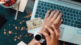 Weibliche Hände über Laptop Tastatur, eine Hand greift an eine Uhr, daneben liegt eine American Express Karte in Gold.