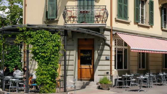 Aussenansicht des Restaurants Olympia in Bern mit grünem Fassadenbewuchs, gemütlicher Terrasse und markanter Holztür. Die rot-weiss gestreifte Markise und der Balkon mit Bistrostühlen runden das charmante Stadtbild ab.