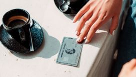 Eine Hand berührt eine silberne Kreditkarte von American Express auf einem hellen Steintisch neben zwei schwarzen Espressotassen im Sonnenschein. 