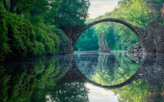 Schmale Bogensteinbrücke, die sich kreisförmig in einem Gewässer spiegelt in einem Waldgebiet. 