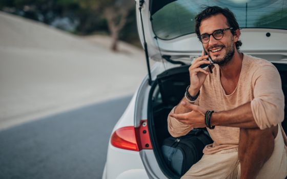 Ein Mann sitzt im Kofferraum eines Mietautos und telefoniert mit seinem Smartphone. 
